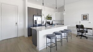 A modern kitchen with a large island, bar stools, a refrigerator, and a dining area with a chair. - Landing | Spectacular 2BD, Pool, Gym (Plano)