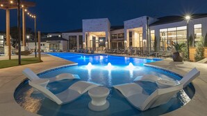 A swimming pool with lounge chairs and a fountain, surrounded by buildings with large windows and outdoor lighting. - Landing | Stunning 1BD, Gym (San Antonio)