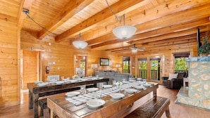 A spacious wooden cabin with a large dining table set for six, a stone fireplace, and a cozy seating area with a couch and a television. - Sleeps 116 | Smoky Mountain Views | 4 Hot Tubs | 2 Game Rooms | (Sevierville)