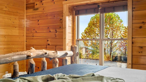 A wooden cabin room with a large window, a wooden railing, and a bed with a green blanket. - Sleeps 116 | Smoky Mountain Views | 4 Hot Tubs | 2 Game Rooms | (Sevierville)