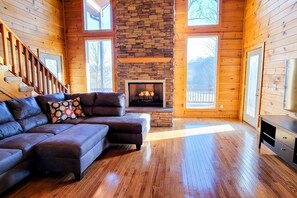 Un acogedor interior de cabaña con chimenea de piedra, pisos de madera, sofá modular y ventanas amplias.