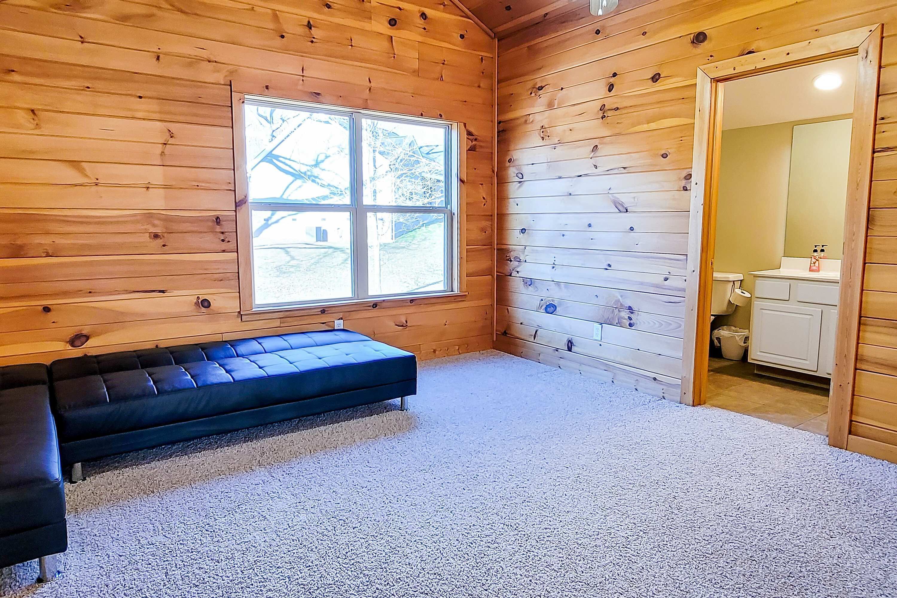 Une pièce aux murs en bois, avec une grande fenêtre, un canapé noir et une salle de bain visible à travers une porte ouverte.