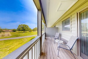 A wooden deck with two chairs and a small table, overlooking a grassy area with trees and a building in the distance. - Inviting & Cozy Studio, Sleep 4, Shared Gym & Games (Baneberry)
