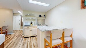 A compact kitchen with white cabinets, a microwave, and a coffee maker on shelves. There is a small island with a sink and two chairs. - Inviting & Cozy Studio, Sleep 4, Shared Gym & Games (Baneberry)