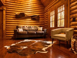A log cabin interior with a leather sofa, a cowhide rug, and a window with autumn leaves. - Mountain Views, Hot Tub & Dog-Friendly Cabin (Gatlinburg)