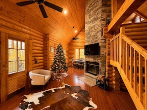 A log cabin interior with a fireplace, a Christmas tree, a sofa, and a stone wall. - Mountain Views, Hot Tub & Dog-Friendly Cabin (Gatlinburg)
