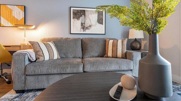 Ein modernes Wohnzimmer mit einem grauen Ecksofa, einer großen Vase mit Grünpflanzen und abstrakter Kunst an der Wand.