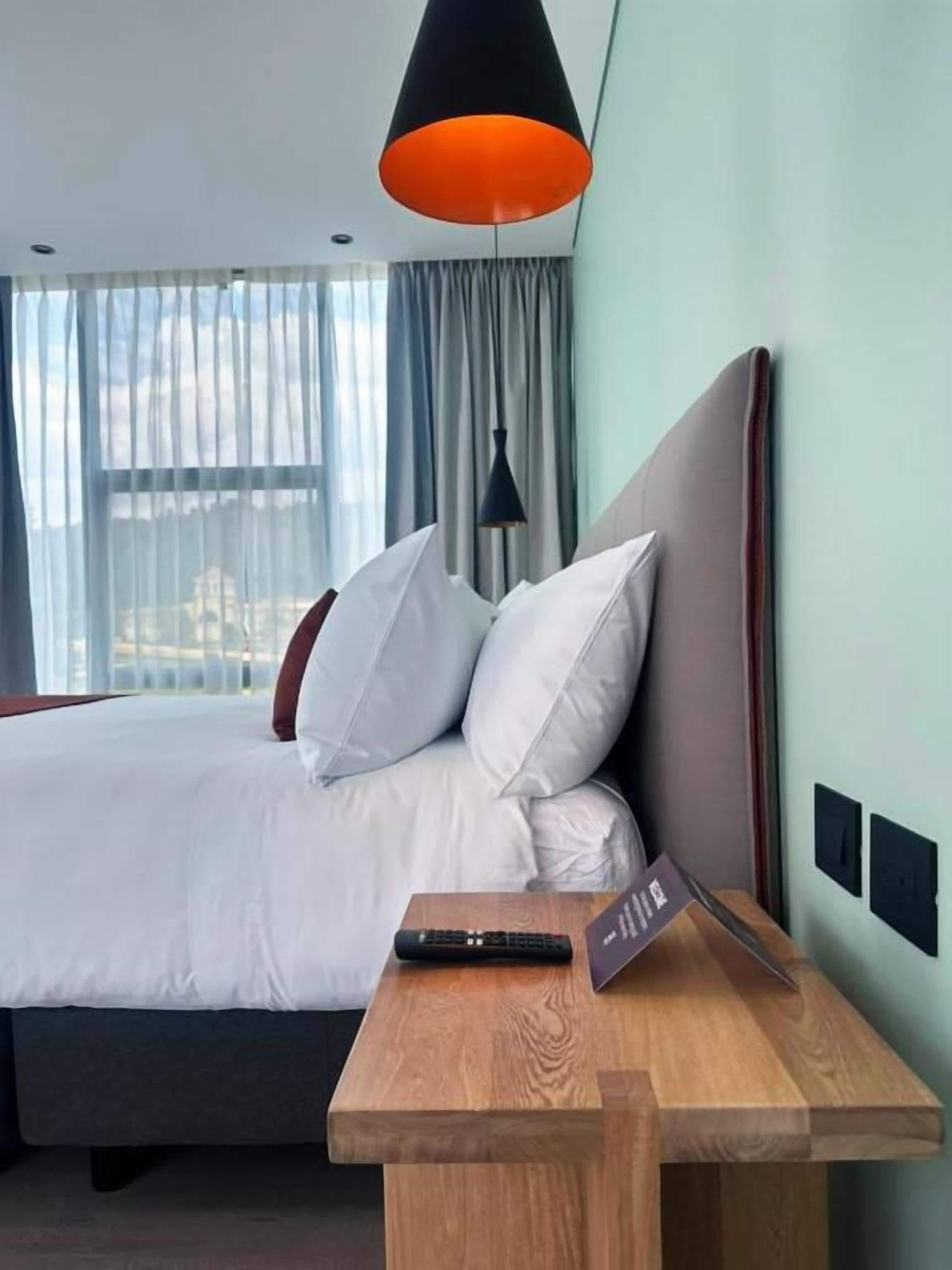 A hotel room with a bed, a wooden nightstand, a remote control, and a lamp.