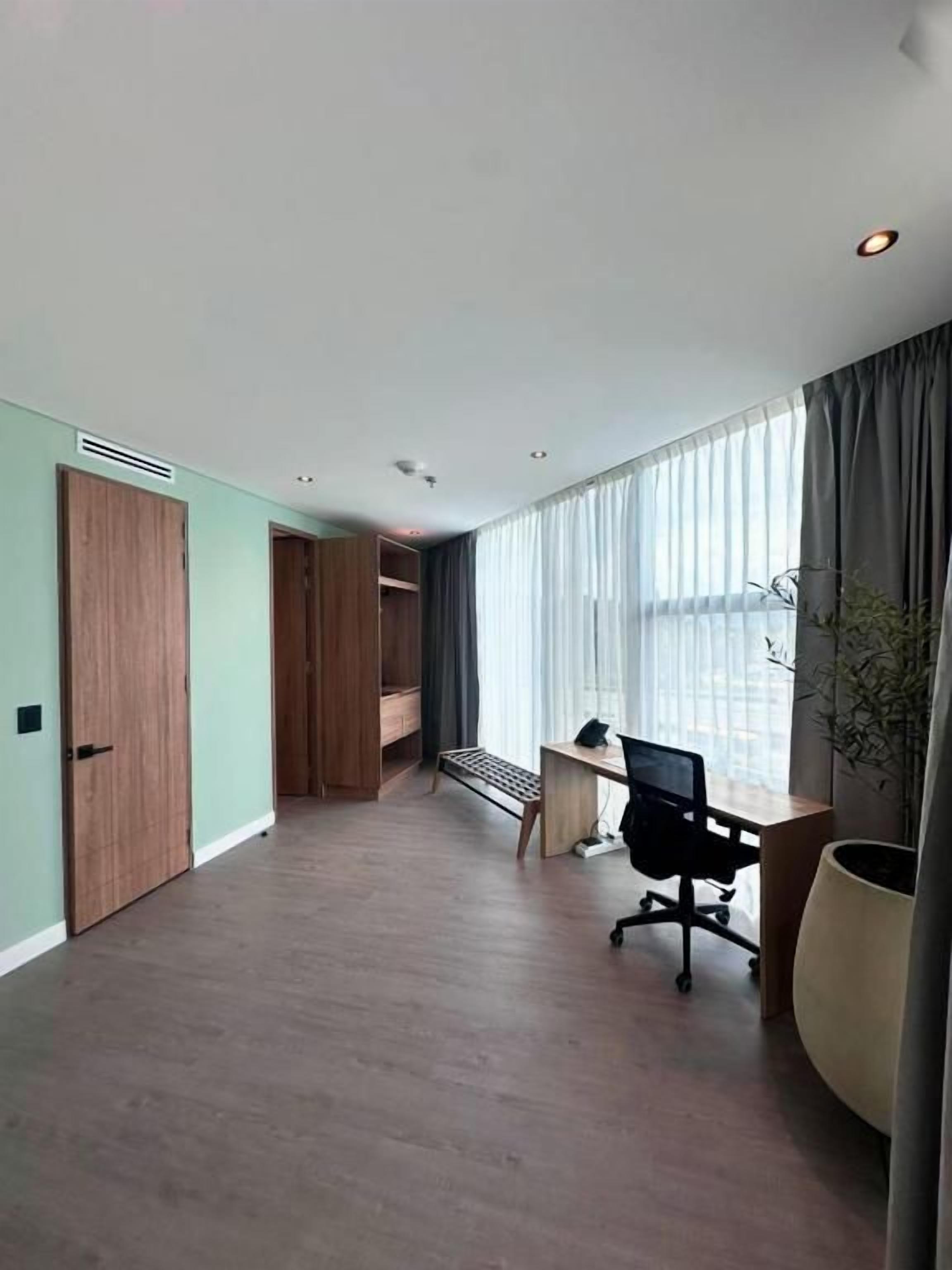 A room with a wooden floor, a desk with a chair, a large window with curtains, and a potted plant.