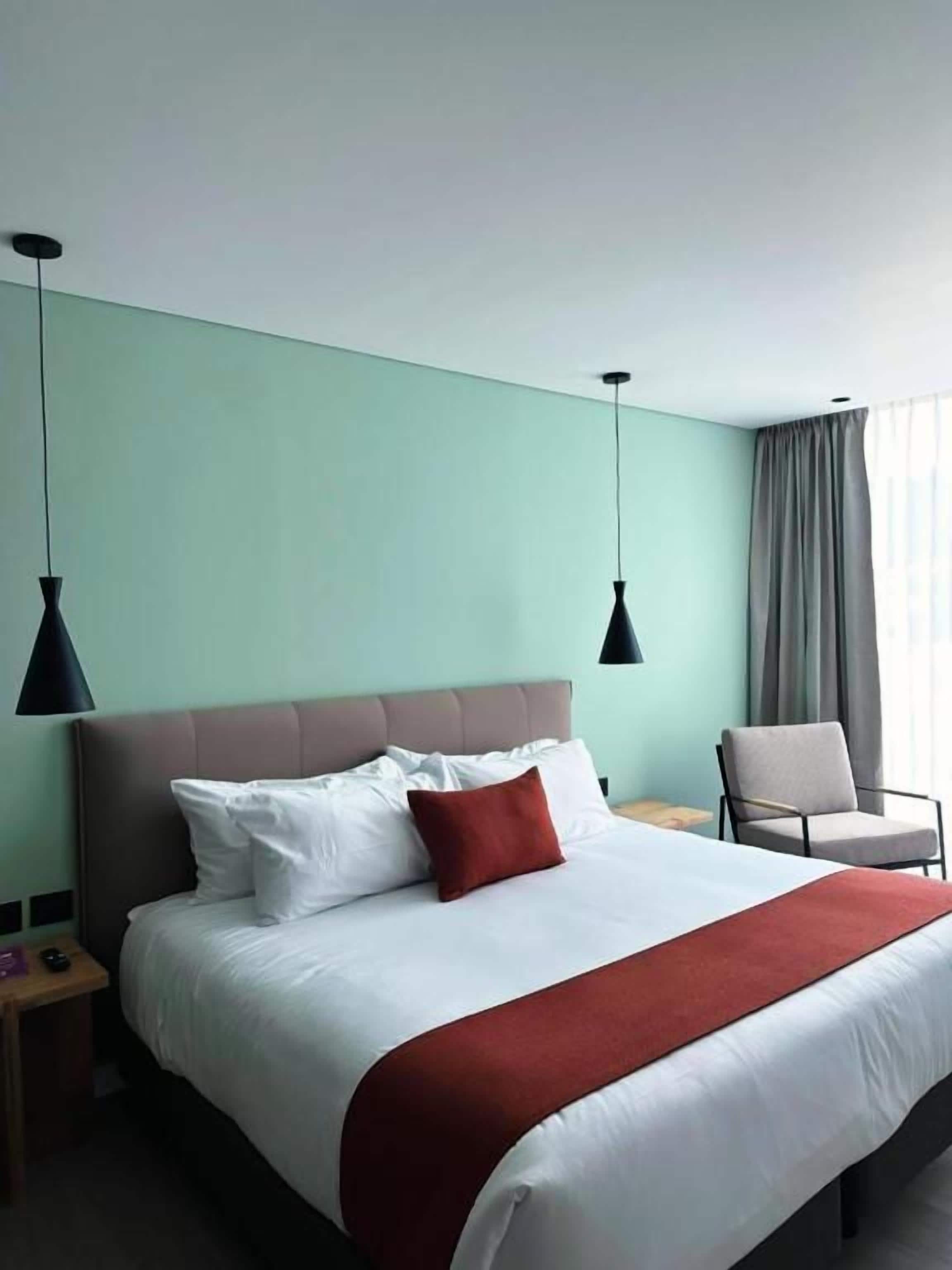 Una habitación de hotel con una cama grande, dos lámparas colgantes, una silla y una mesita de noche.