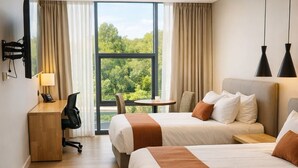 A hotel room with two beds, a desk, a chair, a small table, and a view of greenery through large windows. - Hotel B3 Sibelius (Chía)