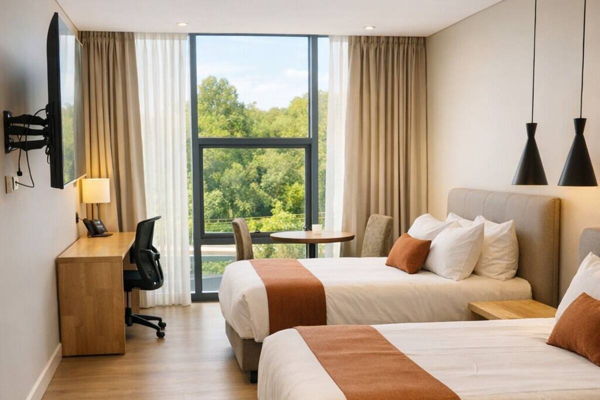 Ein Hotelzimmer mit zwei Betten, einem Schreibtisch, einem Stuhl, einem kleinen Tisch und Blick auf Grün hinter großen Fenstern.