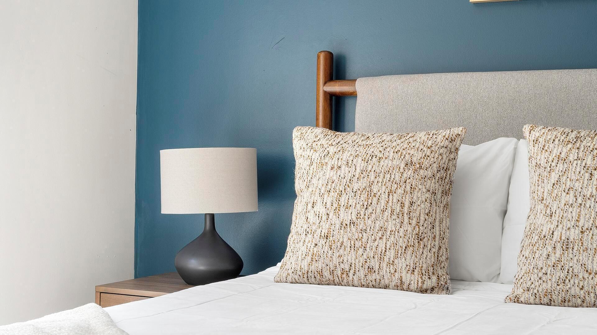 Un lit bien fait, avec une literie blanche, deux coussins à motifs, une lampe de chevet et une tête de lit en bois, le tout adossé à un mur bleu.