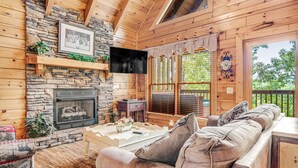 L’intérieur chaleureux d’un chalet avec une cheminée en pierre, des murs en bois, un canapé confortable et une grande fenêtre équipée de stores.