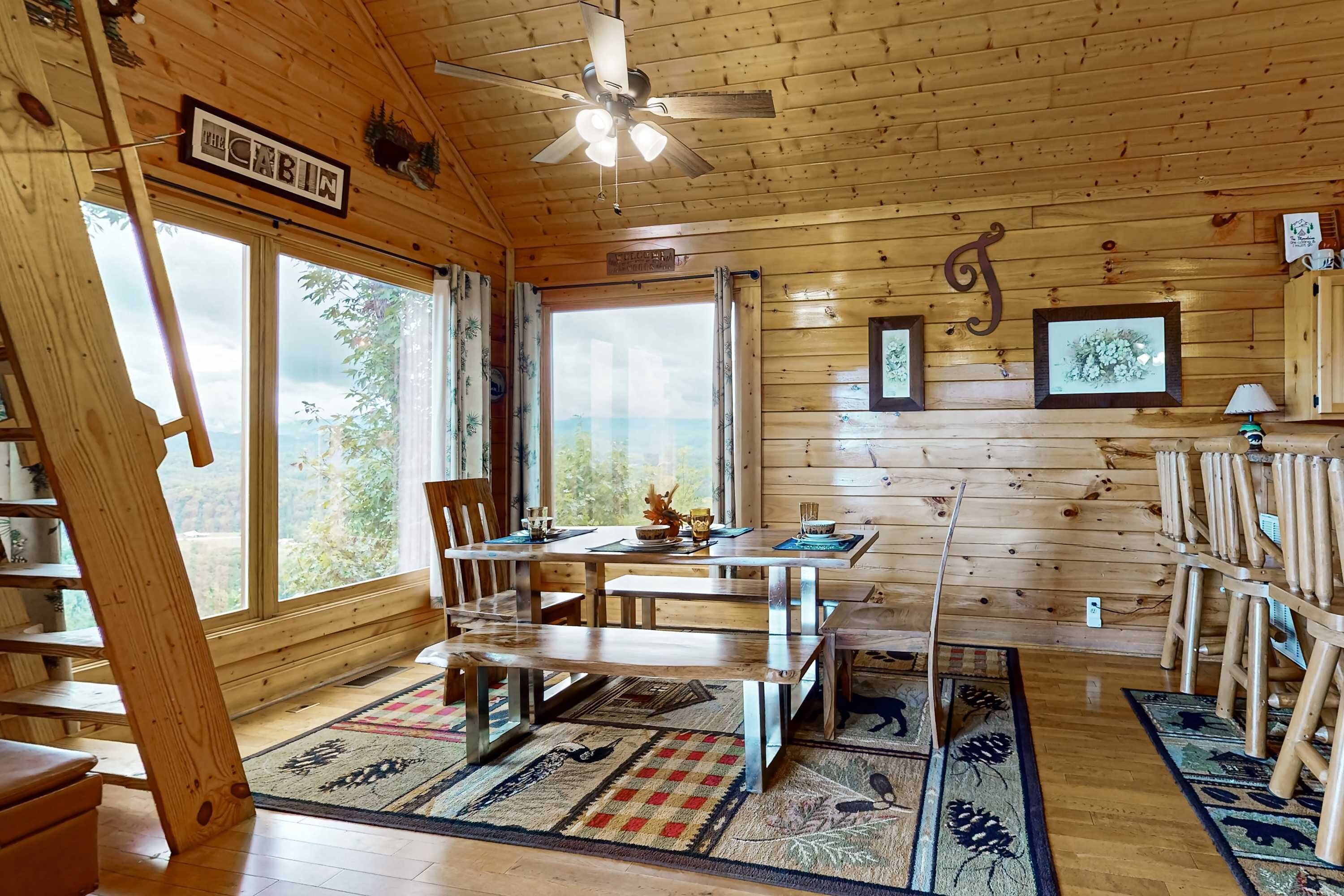 A cabin interior with a wooden dining table, chairs, a ceiling fan, and large windows offering a view of the outdoors.