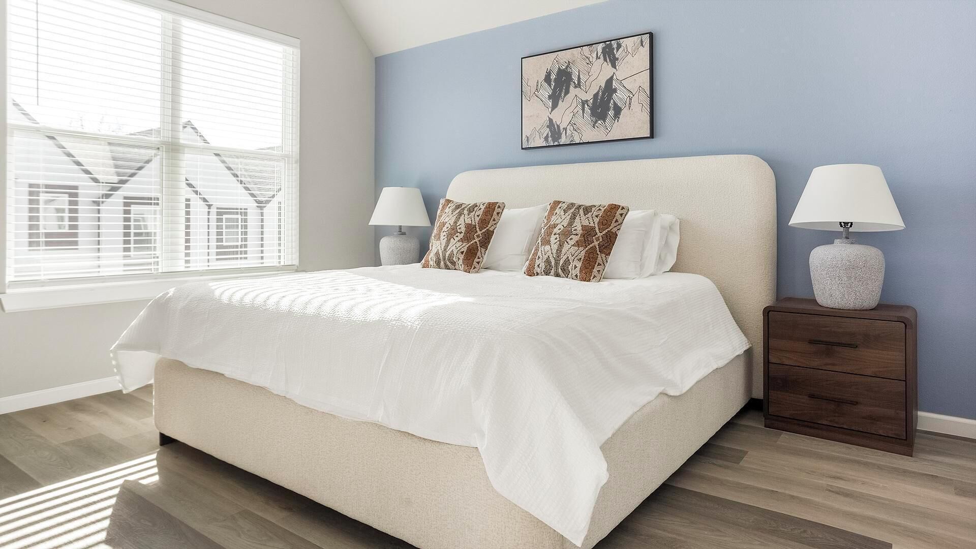 A bedroom with a bed, two bedside tables, a nightstand, a window with blinds, and a wall-mounted picture.