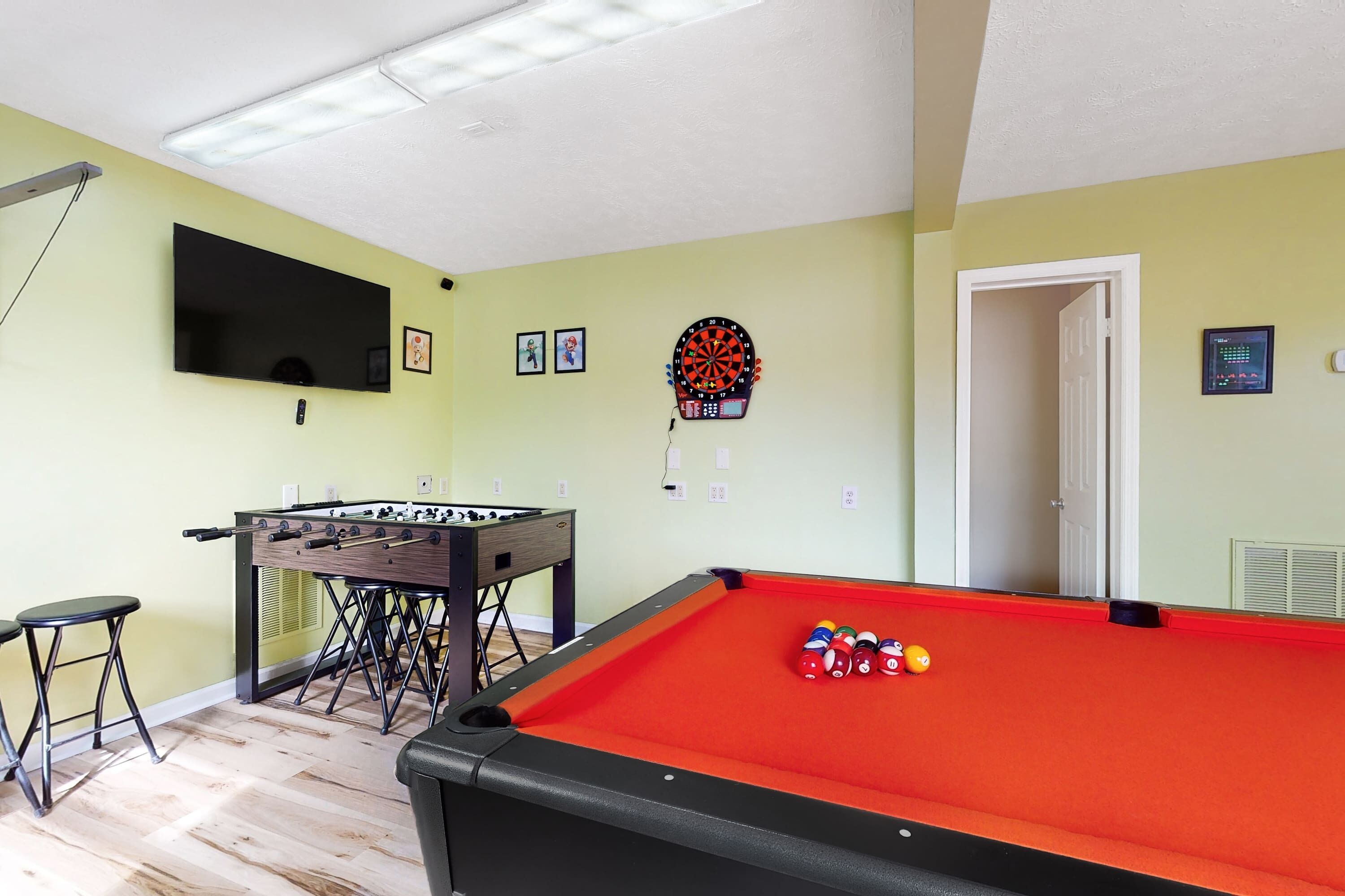 A room with a pool table, foosball table, dartboard, and a television mounted on the wall.