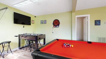 A room with a pool table, foosball table, dartboard, and a television mounted on the wall.