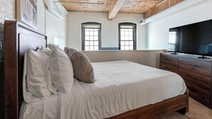 A bedroom with a wooden bed, white bedding, a TV, and a large window. - Landing | Incredible 1BD, Courtyard, Gym (Baltimore)
