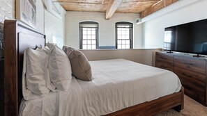 A bedroom with a wooden bed, white bedding, a TV, and a large window. - Landing | Stunning 1BD, Gym, Courtyard (Baltimore)