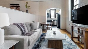A modern living room with a sofa, coffee table, and TV. There is a kitchen area in the background with a dining table and chairs. - Landing | Incredible 1BD, Courtyard, Gym (Baltimore)