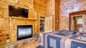 Une chambre rustique en rondins de bois, avec une cheminée, un téléviseur fixé au-dessus et un grand lit recouvert d’une courtepointe.