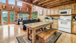 L’intérieur d’un chalet rustique est doté de planchers en bois, d’une table à manger, d’une cuisine avec des armoires en bois, d’une cheminée et d’une grande fenêtre donnant sur une zone boisée.