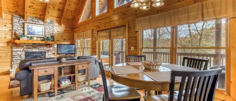 A cozy cabin interior with a stone fireplace, wooden dining table, and large windows offering views of the outdoors.