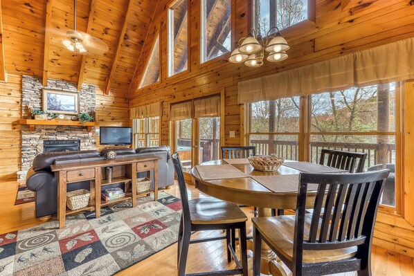 A cozy cabin interior with a stone fireplace, wooden dining table, and large windows offering views of the outdoors. - Mountainside Log Cabin, Sleeps 4, Hot Tub, Game Room (Pigeon Forge)