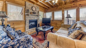 A cozy cabin interior with a stone fireplace, wooden walls, a ceiling fan, and a television. - Family Friendly Luxe Mtn Getaway・Near Downtown (Pigeon Forge)