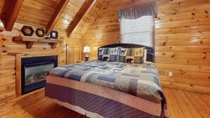 Une chambre confortable dans un chalet, avec un plafond en bois, une cheminée, une couvre-lit matelassé et une fenêtre équipée de stores.