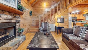 L’intérieur chaleureux d’un chalet avec une cheminée en pierre, un plancher en bois, un canapé, un fauteuil et un escalier menant à un coin cuisine.
