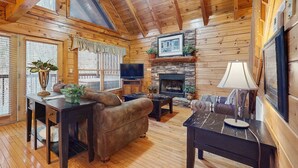 L’intérieur chaleureux d’un chalet avec des murs en bois, une cheminée en pierre, un téléviseur et un coin salon confortable.