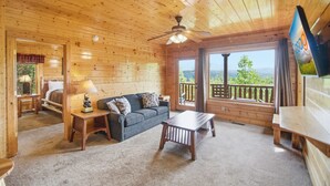 A wooden cabin interior with a sofa, table, ceiling fan, and large windows offering a view. - Cabin W Amazing Views・Hot Tub・Shared Sauna - Pool (Pigeon Forge)