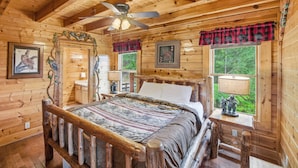 A wooden cabin bedroom with a bed, a ceiling fan, a window with a view, and a lamp on a bedside table. - Cabin W Amazing Views・Hot Tub・Shared Sauna - Pool (Pigeon Forge)