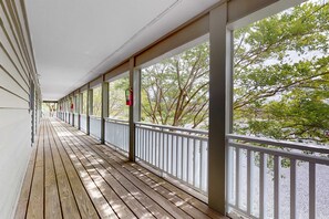 Um deck de madeira coberto com grades brancas e janelas amplas, oferecendo vista para árvores e um caminho de cascalho.