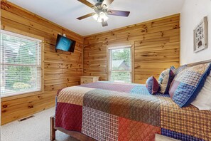 Ein Zimmer mit Holzwänden, einem Bett, einem Deckenventilator, einem an der Wand montierten Fernseher und zwei mit Jalousien versehenen Fenstern.