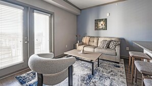 A modern living room with a sofa, coffee table, and chairs. There is a large window with blinds, a rug, and a painting on the wall. - Landing | Stunning 2BD, Gym, Pool (Nashville)