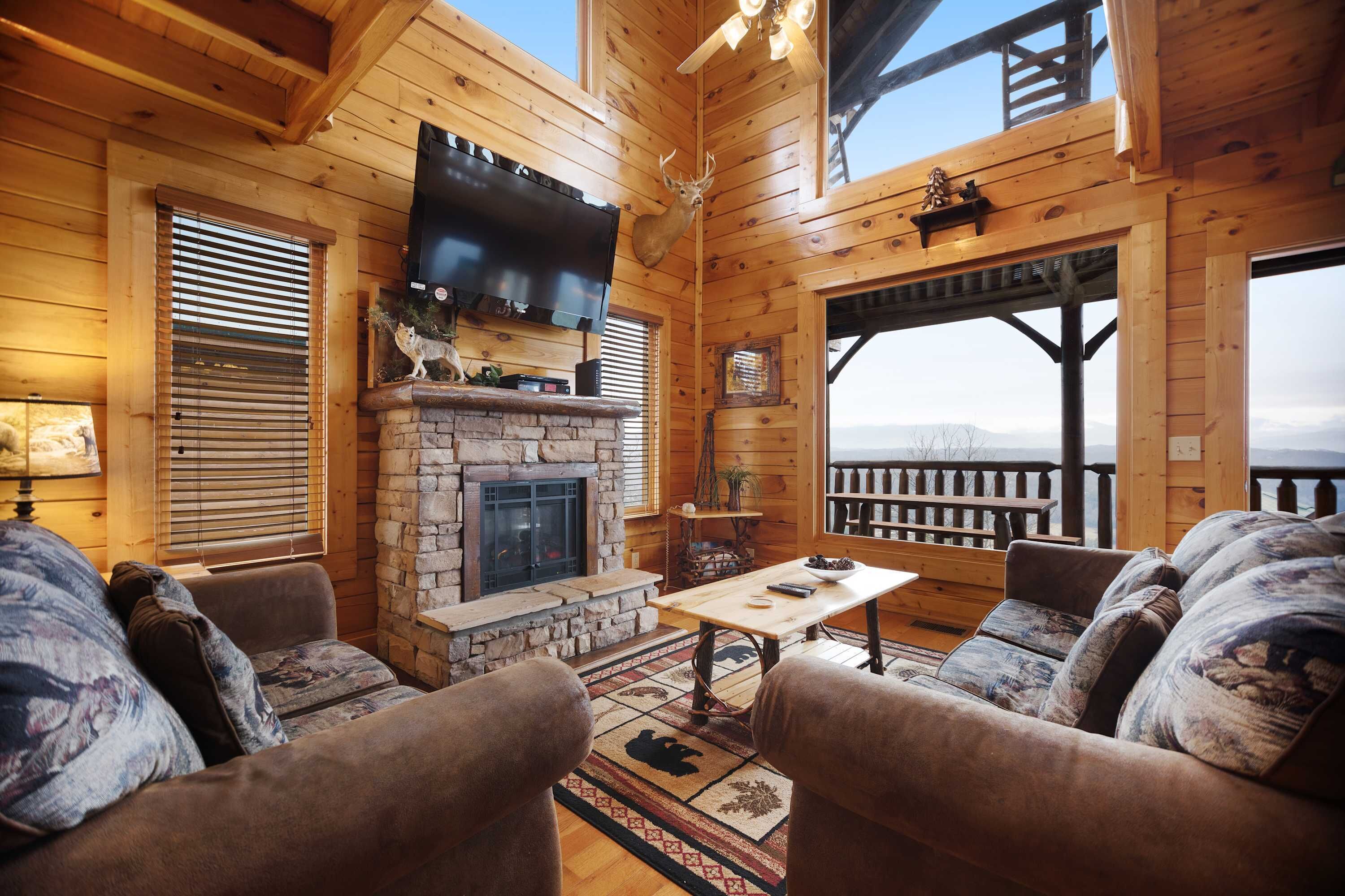 A cozy log cabin interior with a stone fireplace, wooden furniture, and a large TV mounted on the wall.