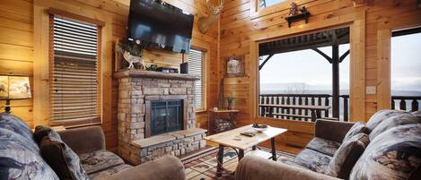 A cozy log cabin interior with a stone fireplace, wooden furniture, and a large TV mounted on the wall.