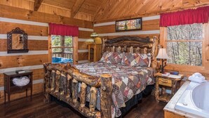 Een slaapkamer in een blokhut met een houten bed, een klein nachtkastje, een venster met uitzicht op bomen en een groot raam met een rode gordijn.