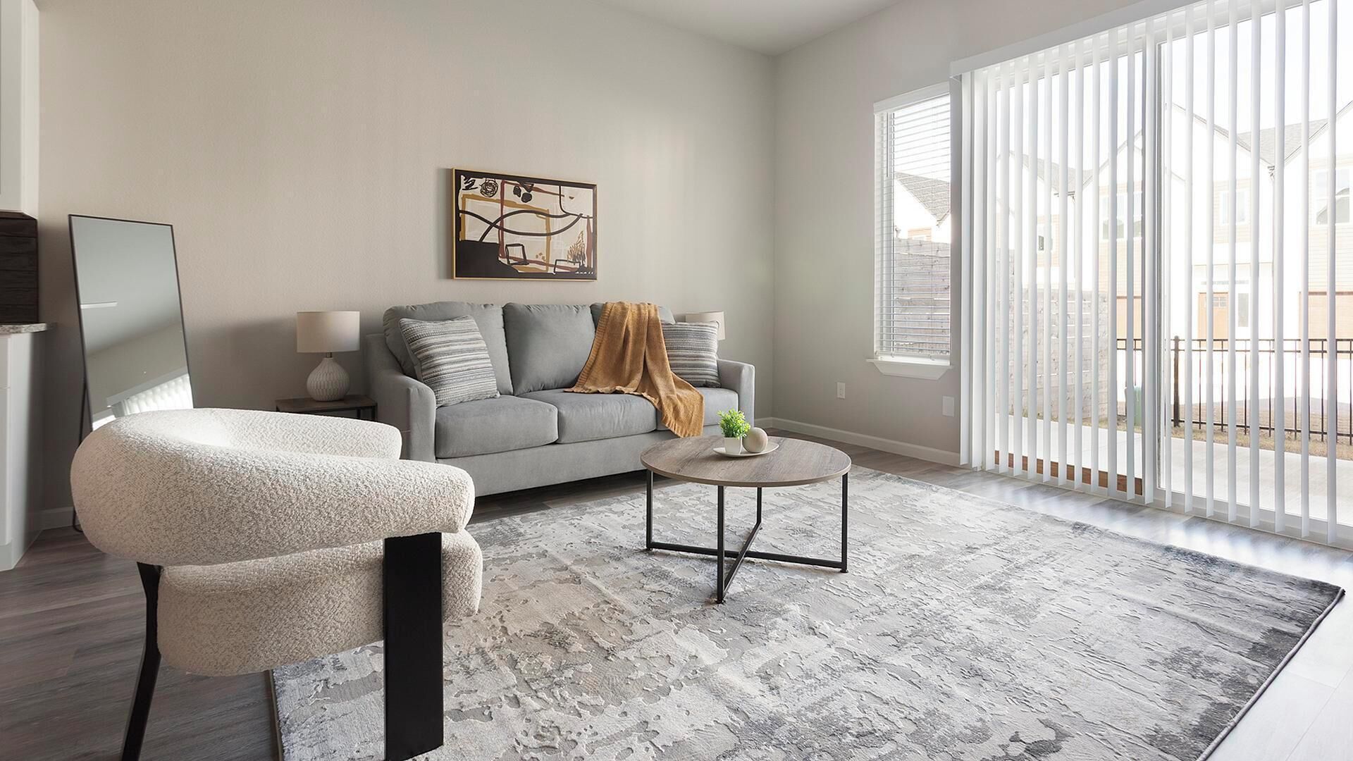A modern living room with a grey sofa, a white armchair, and a round coffee table. There is a painting on the wall and a large window with blinds.