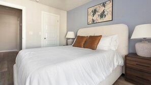 A bedroom with a large bed, two bedside lamps, a door, and a wall-mounted artwork. - Landing | Modern 3BD, Gym, Clubhouse (Sapulpa)