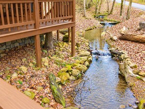 A wooden deck with a stone wall, a small stream, and a forested area with fallen leaves. - Cozy Creekside Cabin Near Pigeon Forge | Sleeps 4 (Pigeon Forge)