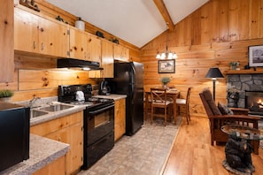 A kitchen with wooden cabinets, a stove, a refrigerator, and a dining area with chairs and a table. - Pigeon Forge Cabin on the Creek | 4 Min to Strip (Pigeon Forge)