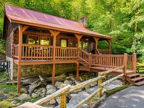 A rustic cabin with a wooden deck and stone foundation, surrounded by lush greenery. - Cozy Creekside Cabin Near Pigeon Forge | Sleeps 4 (Pigeon Forge)
