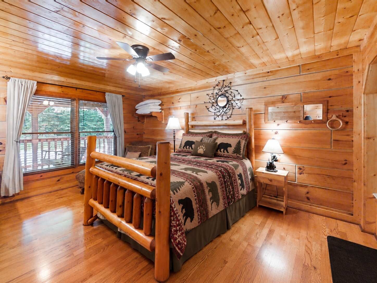 A wooden cabin bedroom with a bed, a ceiling fan, a window with curtains, and a wall-mounted lamp.