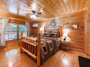 A wooden cabin bedroom with a bed, a ceiling fan, a window with curtains, and a wall-mounted lamp. - Cozy Creekside Cabin Near Pigeon Forge | Sleeps 4 (Pigeon Forge)