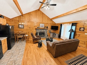 Un intérieur de chalet chaleureux comprenant un coin repas, une cheminée, un téléviseur et un ventilateur de plafond.