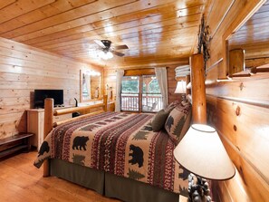 A wooden cabin room with a bed, a television, a window with curtains, and a ceiling fan. - Pigeon Forge Cabin on the Creek | 4 Min to Strip (Pigeon Forge)